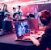 Several people are seated and focused on computer screens, wearing headsets, in an environment that appears to be a gaming tournament or esports event. The room is dimly lit with neon lighting, highlighting screens and creating an intense atmosphere.