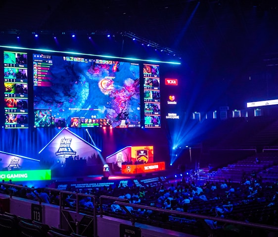 A large esports arena featuring a massive screen displaying a video game, surrounded by sponsor logos and team names. The auditorium is filled with spectators watching the gaming event. Neon blue and purple lights illuminate the stage, creating a vibrant atmosphere.