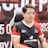 A man with glasses is sitting in a gaming chair, focused on a monitor in front of him. He is wearing a black and red esports jersey with various logos. The background features a large banner with text and numbers in bold fonts.