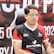 A man with glasses is sitting in a gaming chair, focused on a monitor in front of him. He is wearing a black and red esports jersey with various logos. The background features a large banner with text and numbers in bold fonts.